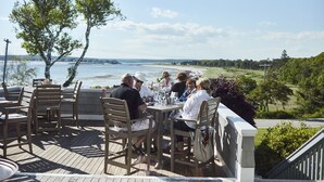 A deck with wooden chairs and tables, overlooking a beach and water.