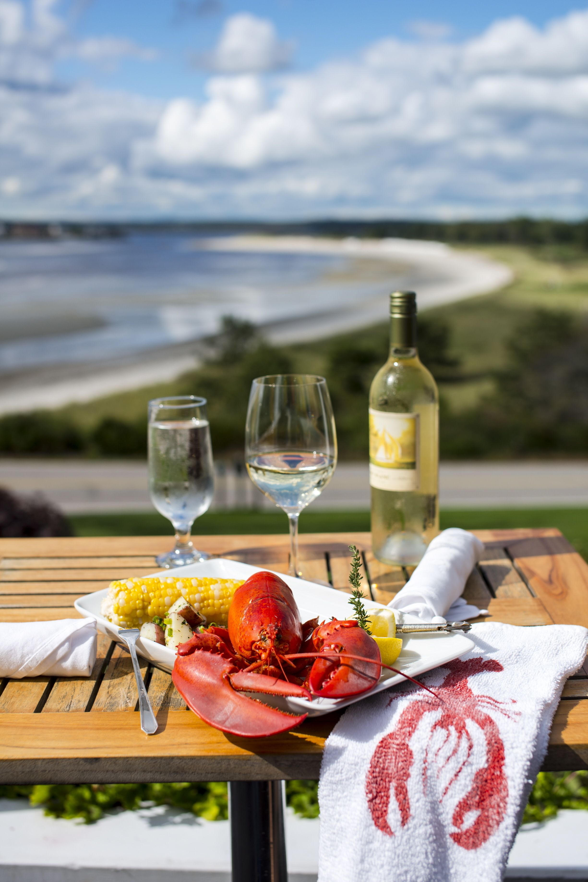 A table with lobster, corn, and wine glasses, set against a scenic beach view.