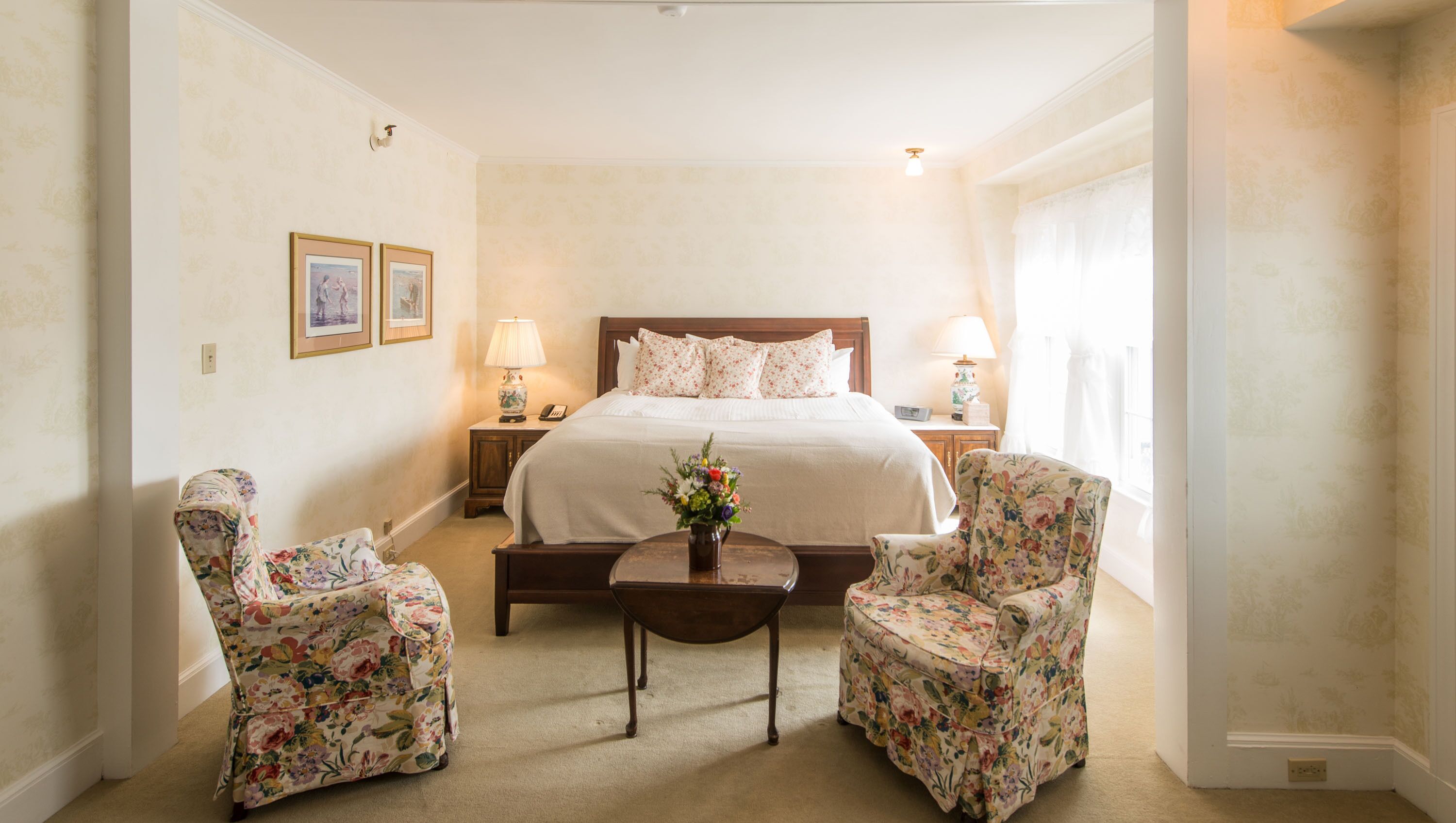 A hotel room with a large bed, two floral armchairs, a small table with a vase, and a nightstand with a lamp.