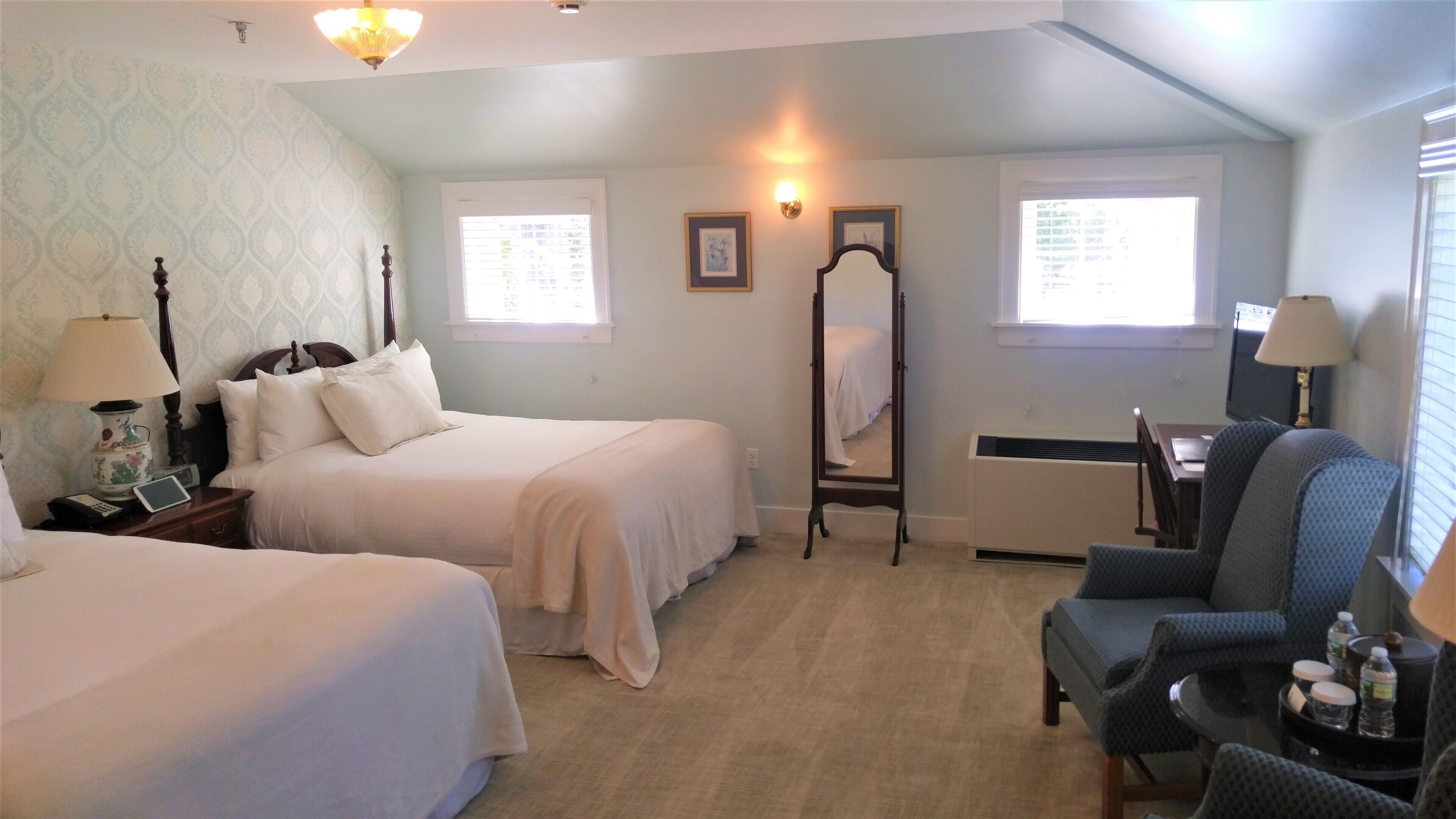 A hotel room with two beds, a desk, a chair, a mirror, and a lamp.