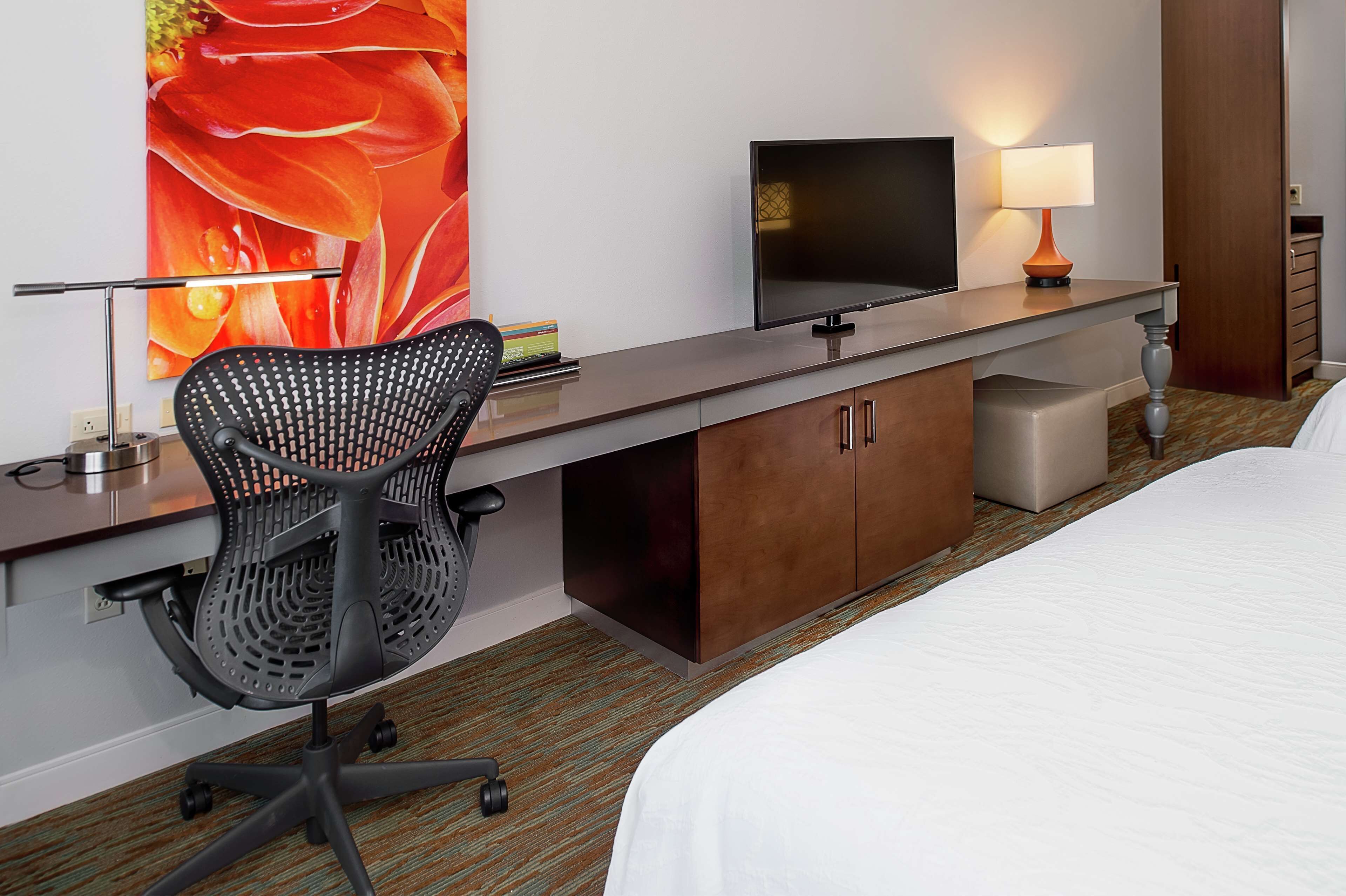 Habitación de hotel con una cama, un escritorio con monitor de computadora, una silla, una lámpara y una gran obra de arte floral en la pared.