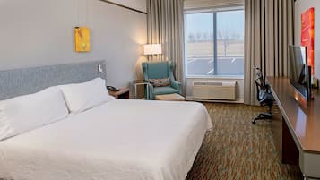 A hotel room with a large bed, a chair, a desk with a TV, a lamp, and a window with a view of a parking lot.