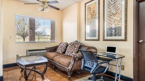 A room with a brown sofa, a glass-top coffee table, a black office chair, a laptop on a desk, and a ceiling fan. - Econo Lodge  Inn & Suites Macon West (Macon)
