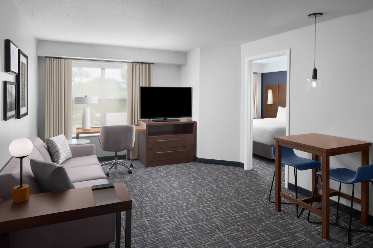 A hotel room with a flat-screen TV, a desk with two chairs, a sofa, a bed, and a large window with curtains.