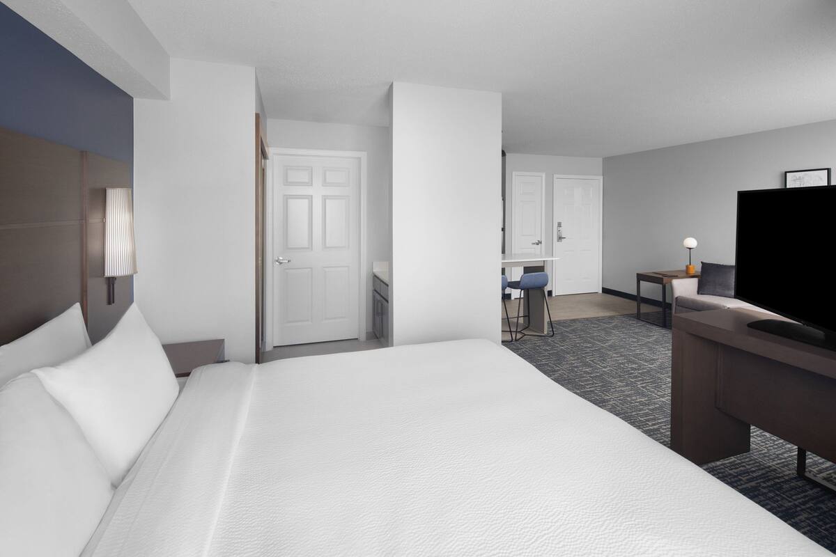 A hotel room with a large bed, a desk with a chair, a television, and a door leading to another room.