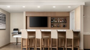 Een moderne bar met een wit aanrecht, vier barkrukken, een flatscreen televisie aan de muur en een plank met verschillende flessen.