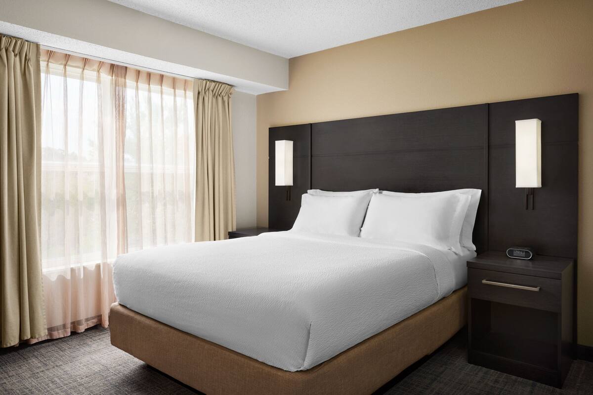 A hotel room with a large bed, a nightstand, a lamp, and sheer curtains.