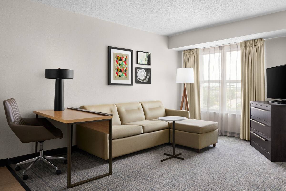 A hotel room with a sofa, a desk, a chair, a small round table, a TV, and two framed pictures on the wall.