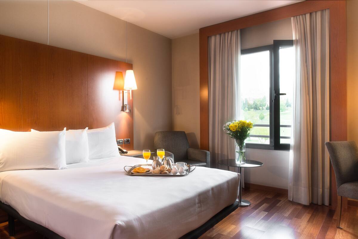 A hotel room with a large bed, a bedside table with a lamp, a chair, a small table with a vase of flowers, and a window with curtains.