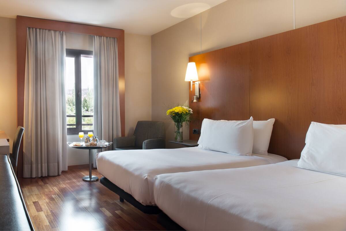 A hotel room with two beds, a small table with breakfast, a chair, a lamp, and a window with curtains.