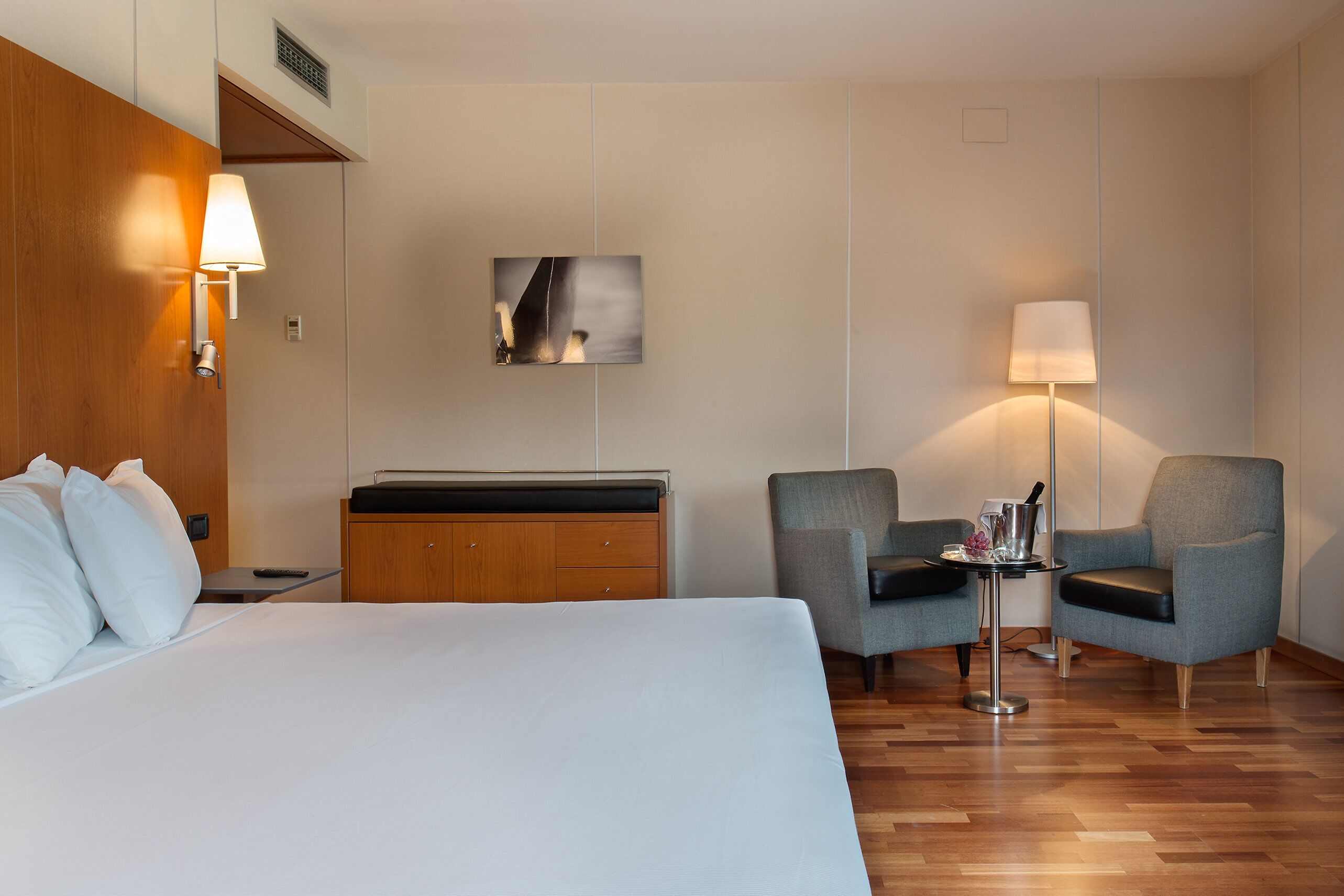 Un cuarto de hotel con una cama grande, dos sillones, una mesita, una lámpara, un cuadro en la pared y un armario de madera.