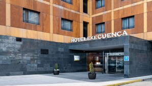 L’entrée d’un hôtel, avec une façade moderne, de grandes fenêtres et une enseigne indiquant « HÔTEL EXECUENCIA ».
