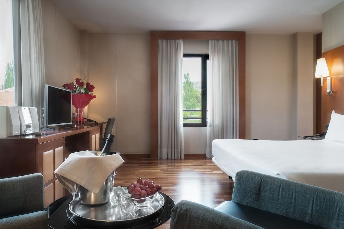 Una habitación de hotel con una cama, un escritorio con una computadora, una silla, una bandeja con un cuenco de fruta y una ventana con cortinas.