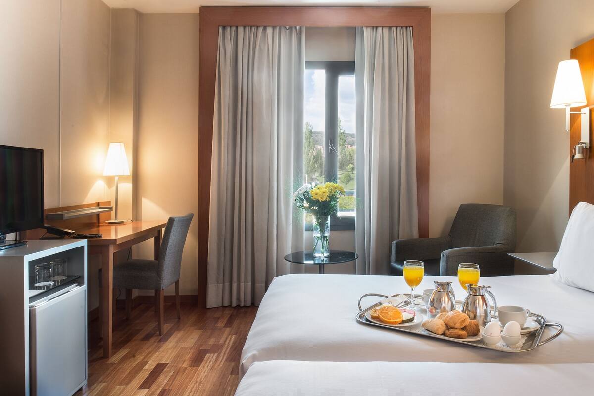 A hotel room with a bed, a desk, a TV, a mini-fridge, a breakfast tray with eggs, juice, and pastries, and a window with curtains.