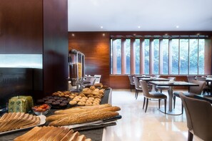 Ein Frühstücksbuffet mit verschiedenen Backwaren und Brotsorten, einer Kaffeestation sowie einem Sitzbereich mit Tischen und Stühlen.