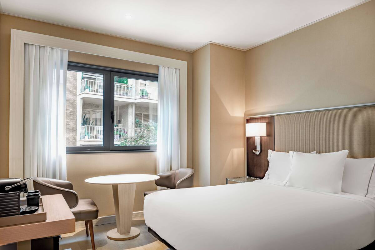 A hotel room with a bed, a desk, a chair, a round table, and a window with curtains.
