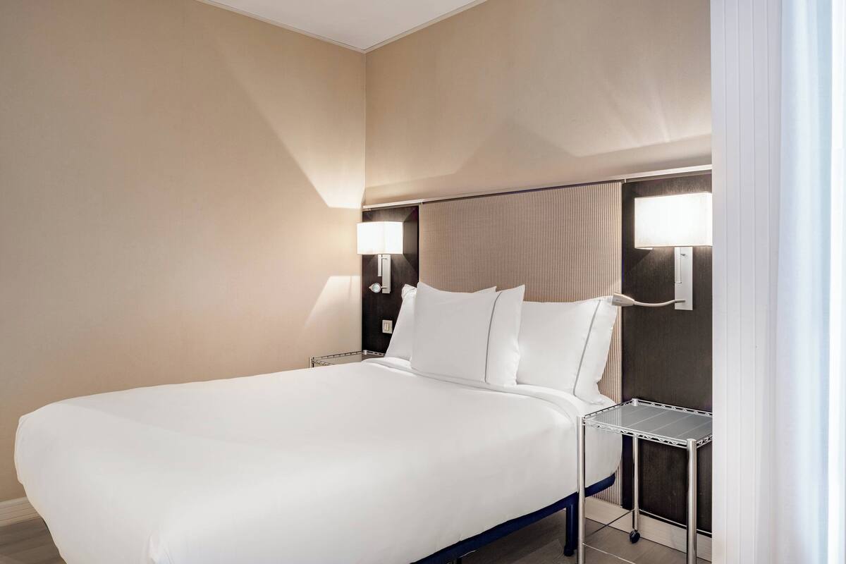 A large bed with white linens, two bedside tables with lamps, and a neutral-colored wall.