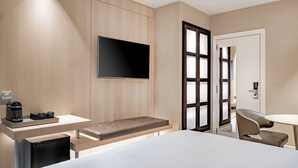 A modern hotel room with a bed, a TV mounted on the wall, a small desk with a coffee maker, and a seating area with a bench and chairs. - AC Hotel Irla by Marriott (Barcelona)