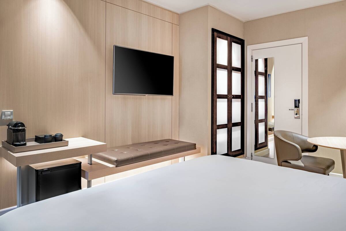 A modern hotel room with a bed, a TV mounted on the wall, a small desk with a coffee maker, and a seating area with a bench and chairs.