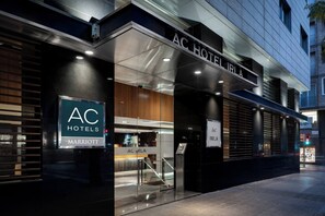 Una entrada de hotel moderna con fachada de vidrio, un mostrador de recepción a la vista y un letrero que indica que forma parte de la cadena AC Hotels Marriott.