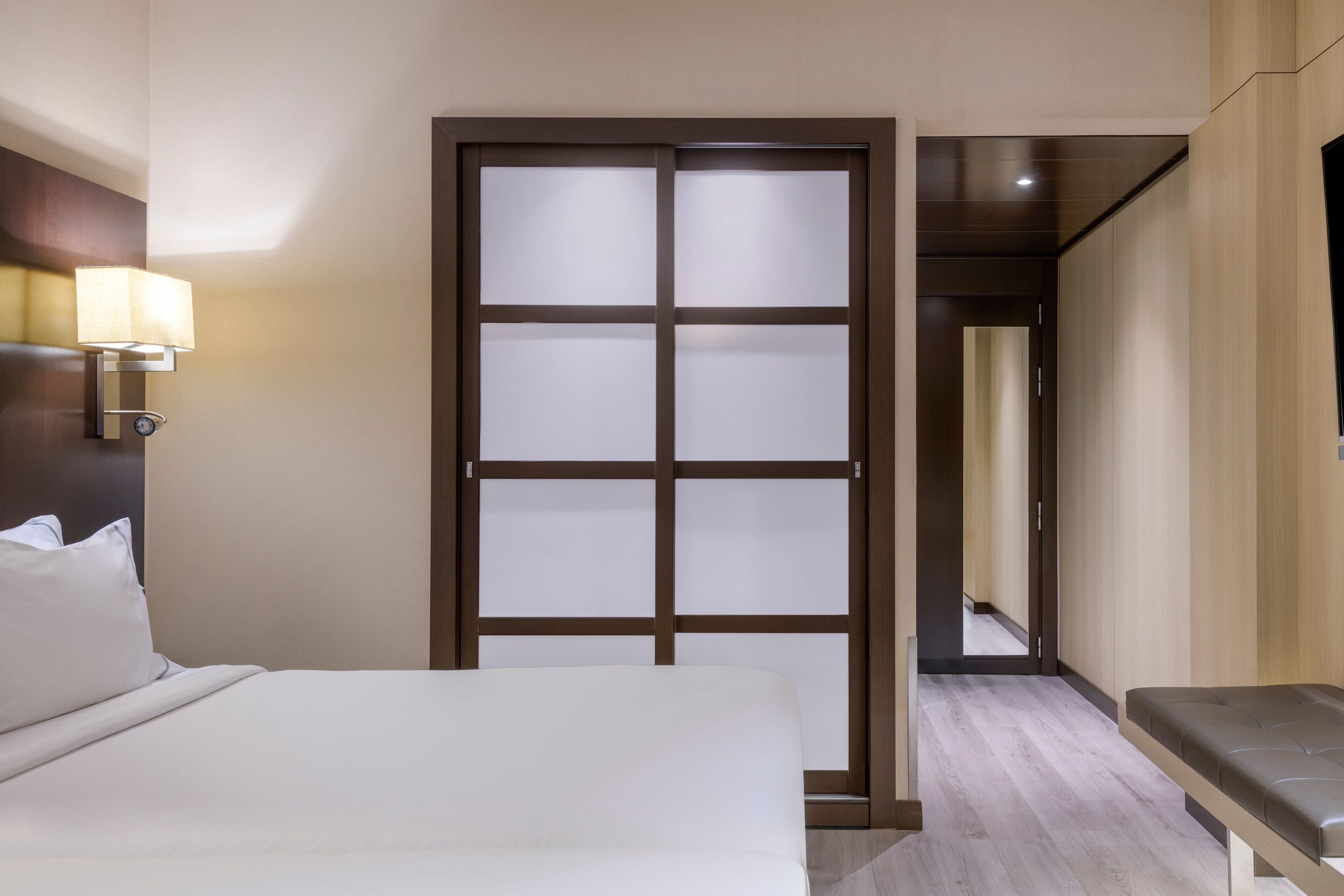 Habitació d'hotel amb llit, una banca, una làmpada de paret i una porta corredissa.