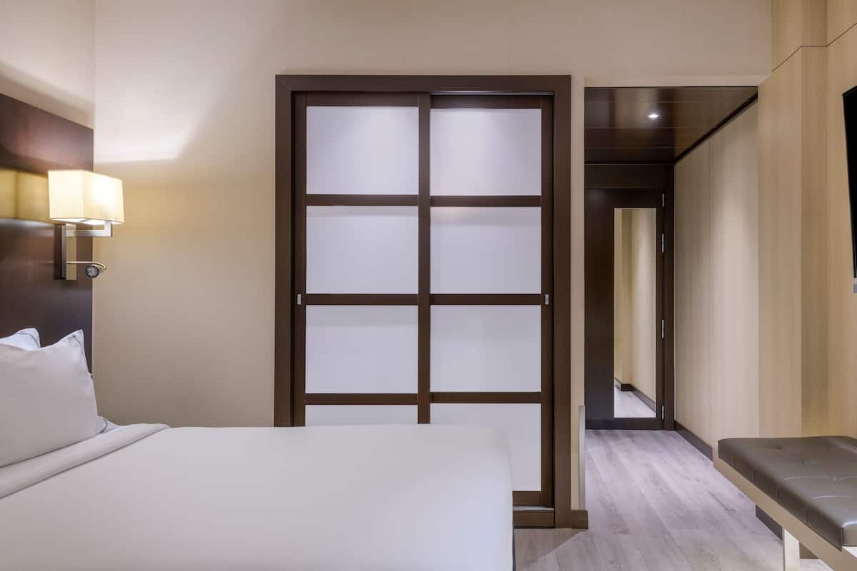 Een hotelkamer met een bed, een bankje, een wandlamp en een matglazen deur.