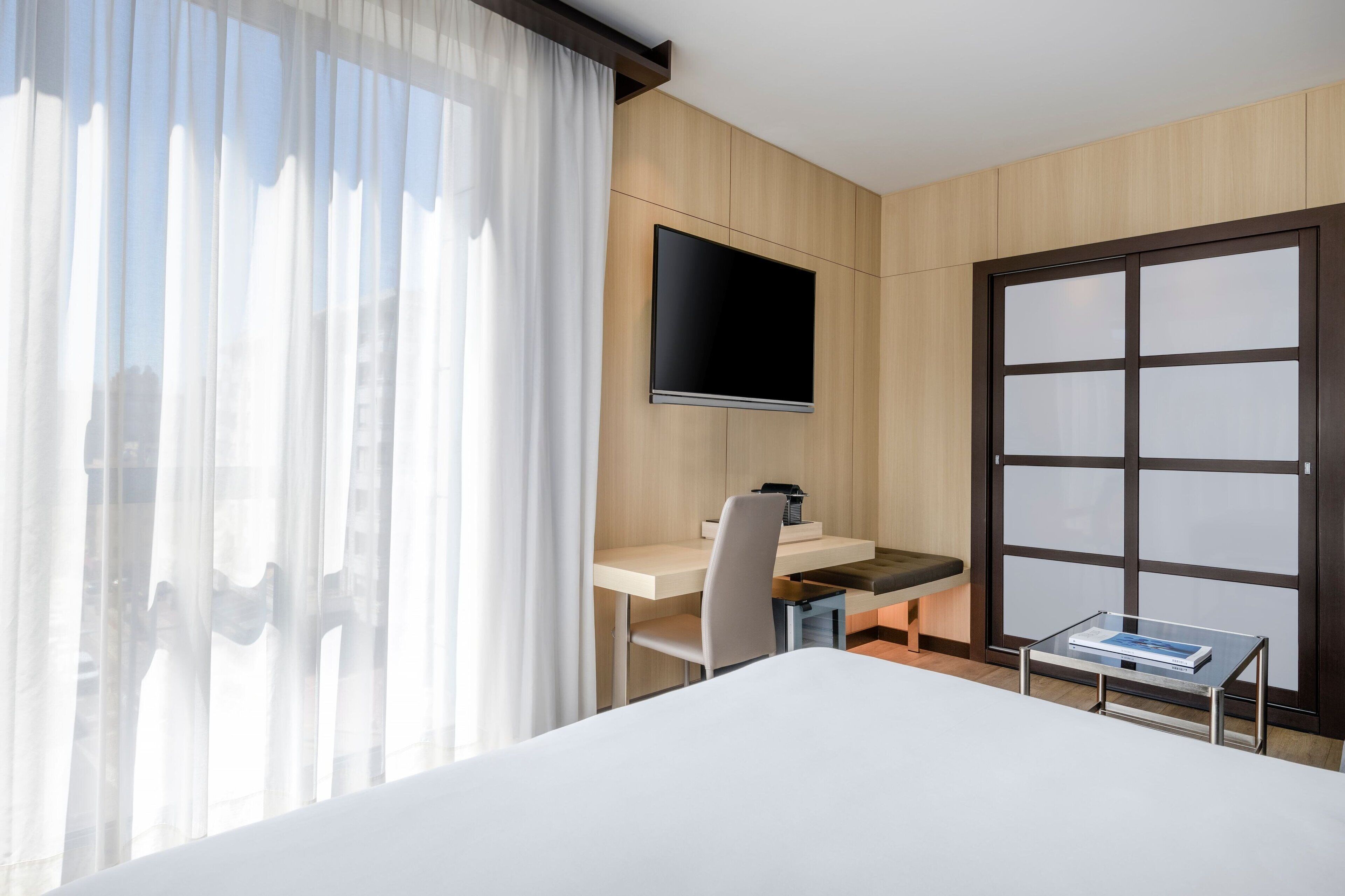 A hotel room with a bed, a desk with a chair, a television, and a window with curtains.
