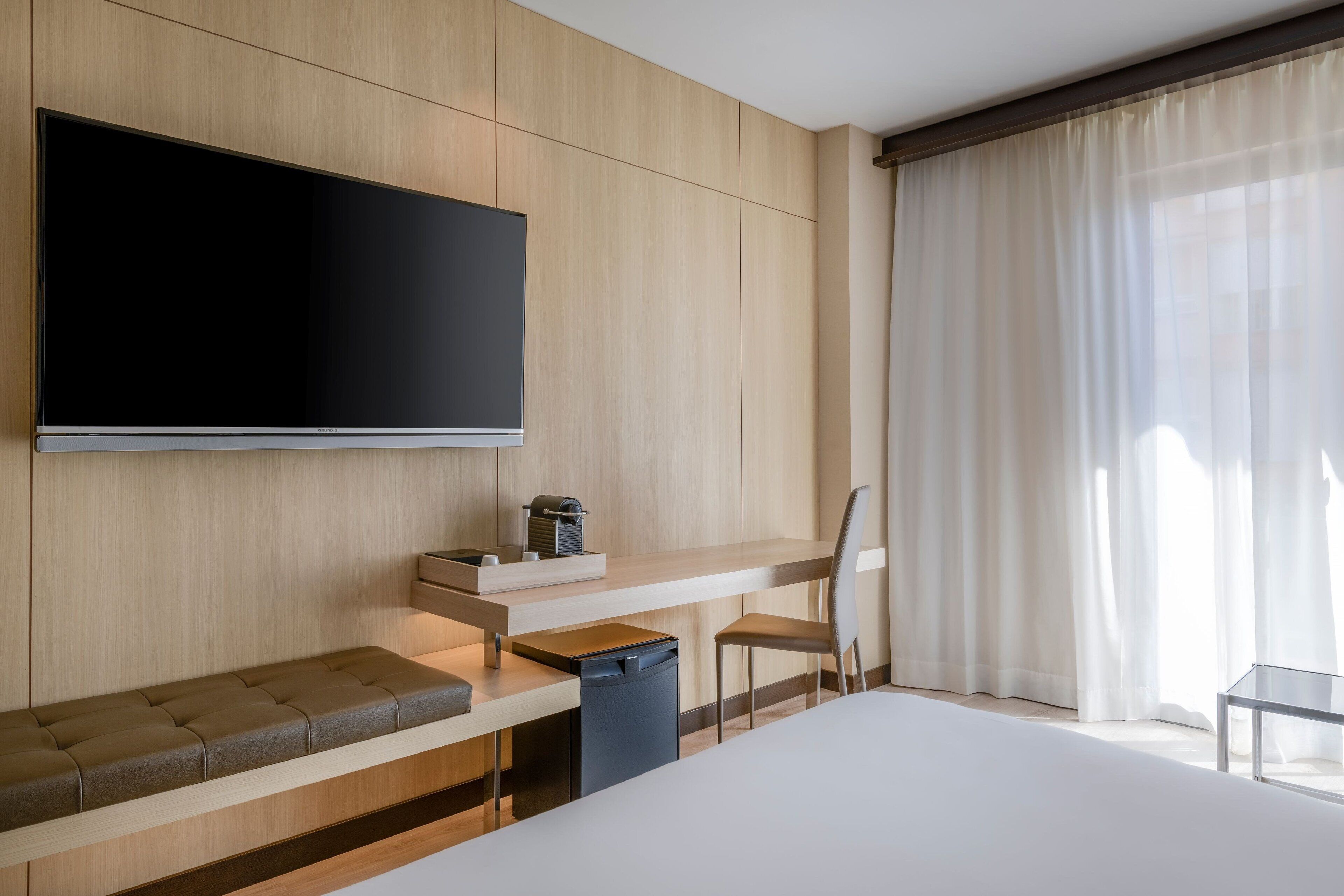 A modern hotel room with a large bed, a flat-screen TV mounted on the wall, a desk with a chair, and a window with sheer curtains.