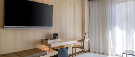 A modern hotel room with a large bed, a flat-screen TV mounted on the wall, a desk with a chair, and a window with sheer curtains.