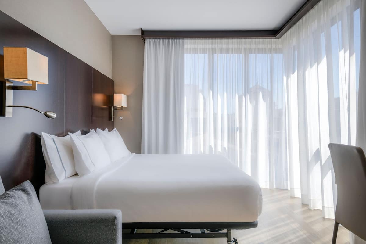 Ein Hotelzimmer mit einem großen Bett, einem Schreibtisch und einem Stuhl. Zwei Wandlampen und ein Fenster mit transparenten Vorhängen sind vorhanden.