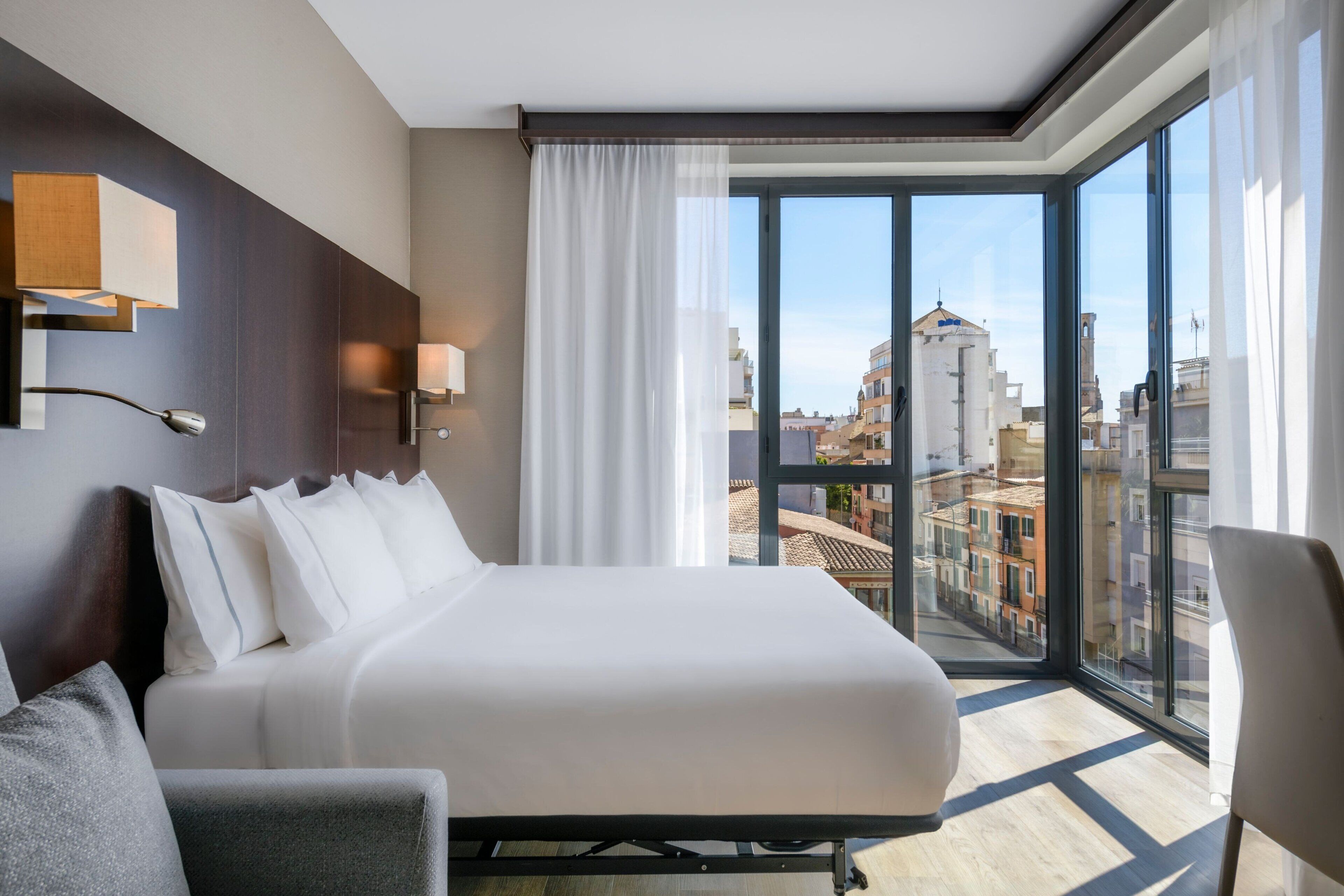 Habitación de hotel con una cama grande, un escritorio y vistas de la ciudad a través de ventanas de pared a techo.