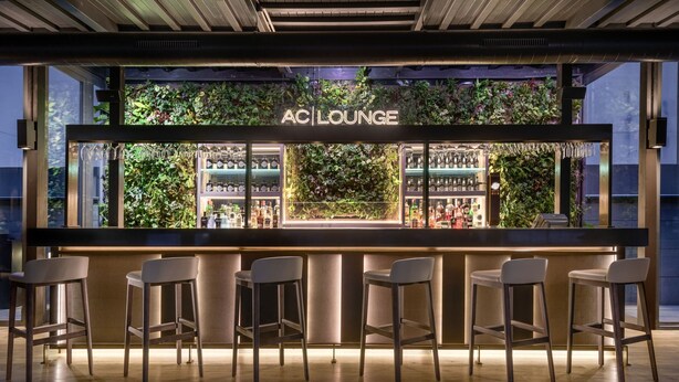 A bar area with a neon sign reading "AC LOUNGE," surrounded by bar stools and a wall of greenery.