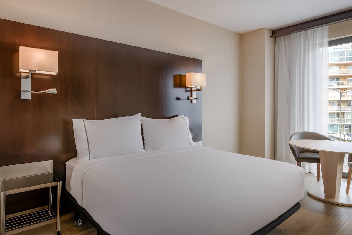 Een hotelkamer met een groot bed, twee wandlampen, een klein tafeltje en uitzicht op een gebouw door het raam.