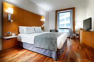 A hotel room with a wooden headboard, a bed with white linens, a desk with a chair, a television, and a window with a view of a building.