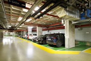 Ένα πάρκινγκ με πολλά αυτοκίνητα σταθμευμένα σε καθορισμένες θέσεις, μια πινακίδα σήμανσης που υποδεικνύει την έξοδο και εμφανή δομικά στοιχεία, όπως σωλήνες και κολώνες από σκυρόδεμα.