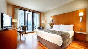 Habitación de hotel con una cama grande, un escritorio con televisor, una silla, una lámpara de escritorio, una mesita de noche con un florero y vistas de la ciudad a través de la ventana.