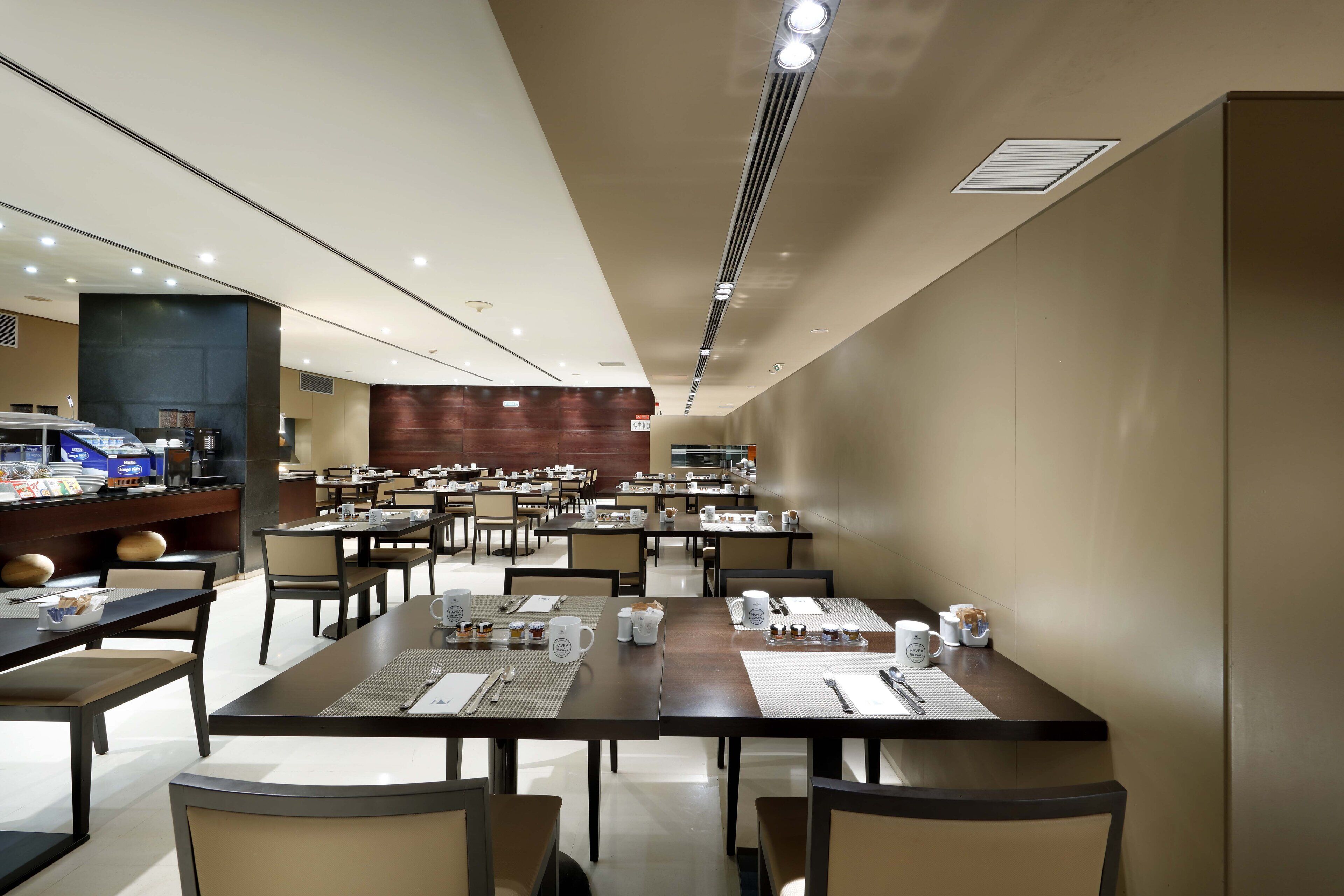 A spacious dining area with multiple tables and chairs, a counter with a coffee machine, and a modern ceiling design.