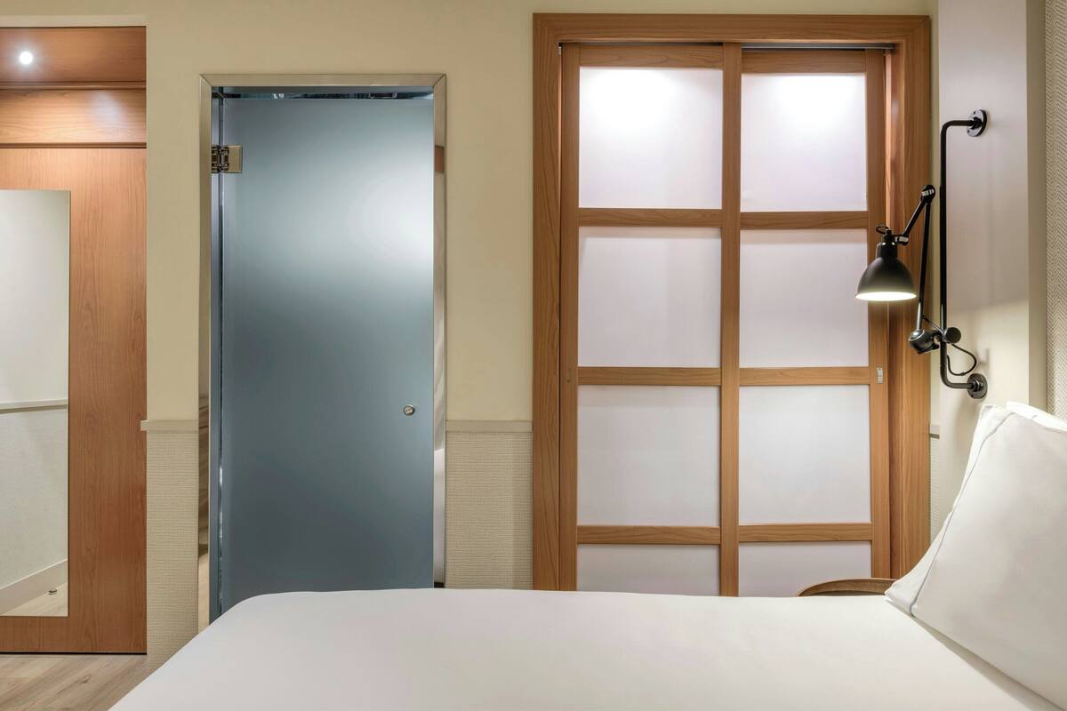 Habitación de hotel con cama, puerta de madera, puerta de vidrio satinado y una lámpara fijada a la pared.