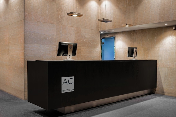 A reception desk with a sign reading "AC HOTELS" in a modern lobby with marble walls and recessed lighting.