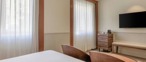 Habitación de hotel con cama, escritorio, silla, televisor montado en la pared y ventana con cortinas.