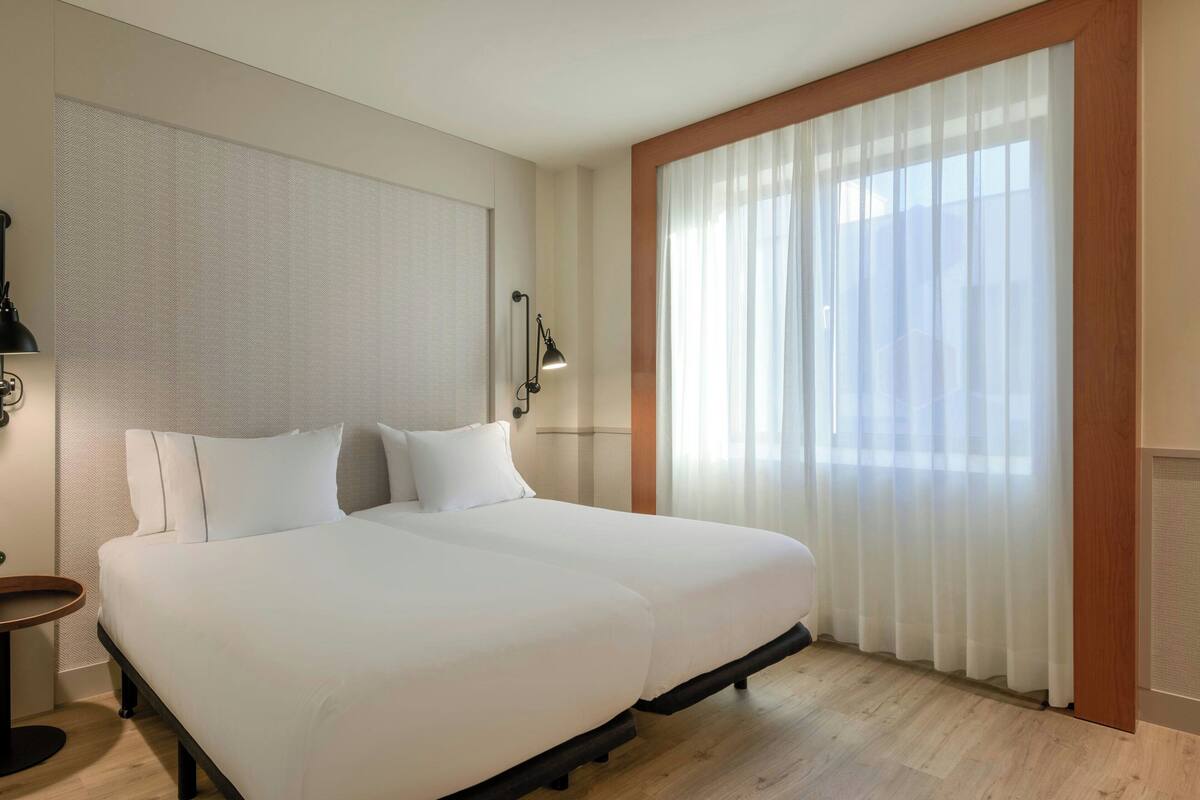 Habitación de hotel con una cama grande, una mesita de noche, una lámpara y una ventana con cortinas de gasa.