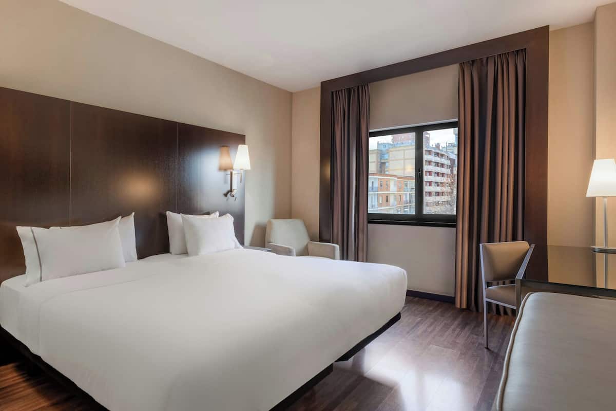A hotel room with a large bed, two bedside tables with lamps, a chair, a window with curtains, and a view of buildings outside.
