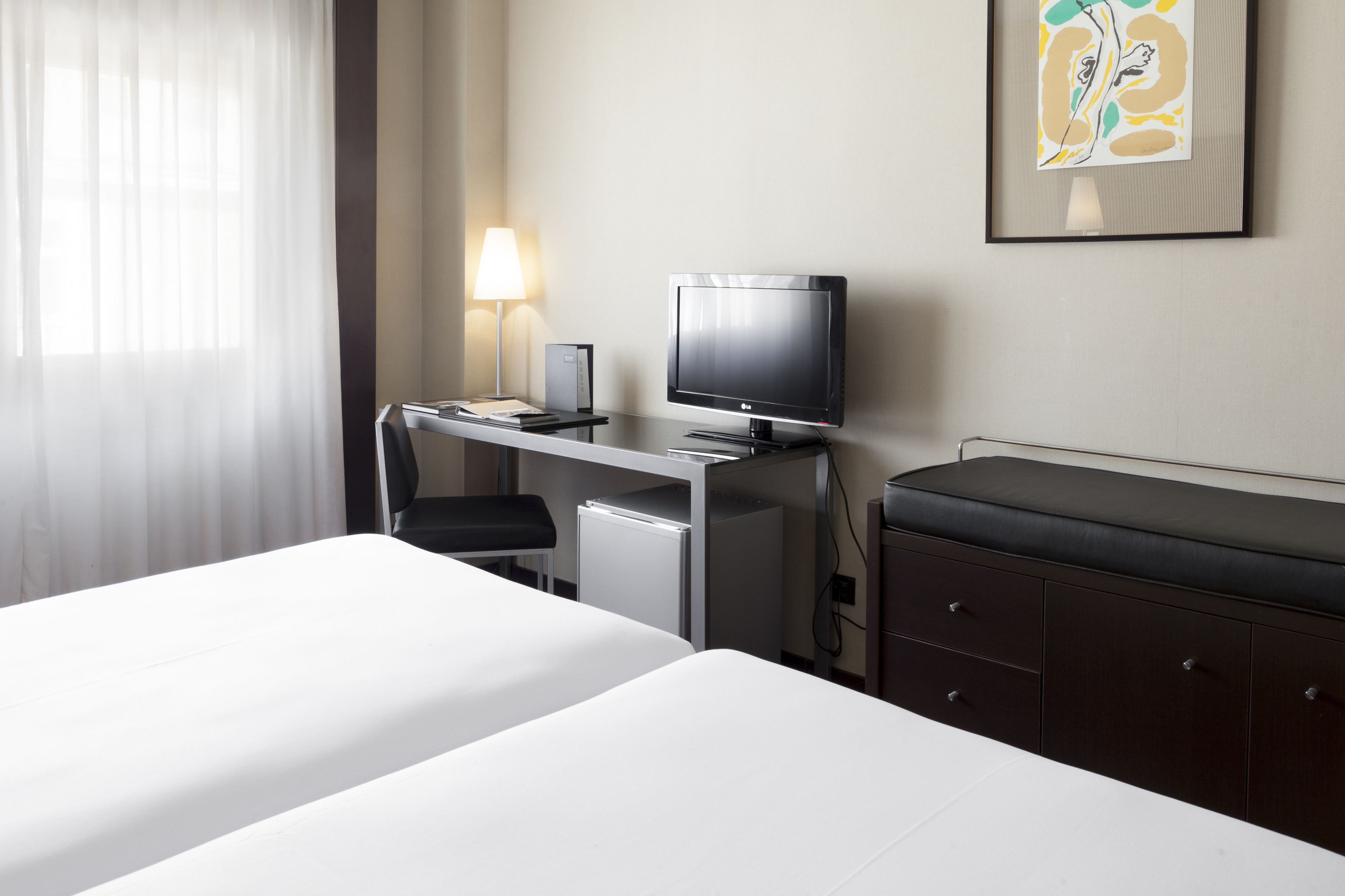 Una habitación de hotel con cama, escritorio con ordenador y lámpara, un refrigerador pequeño y un armario.