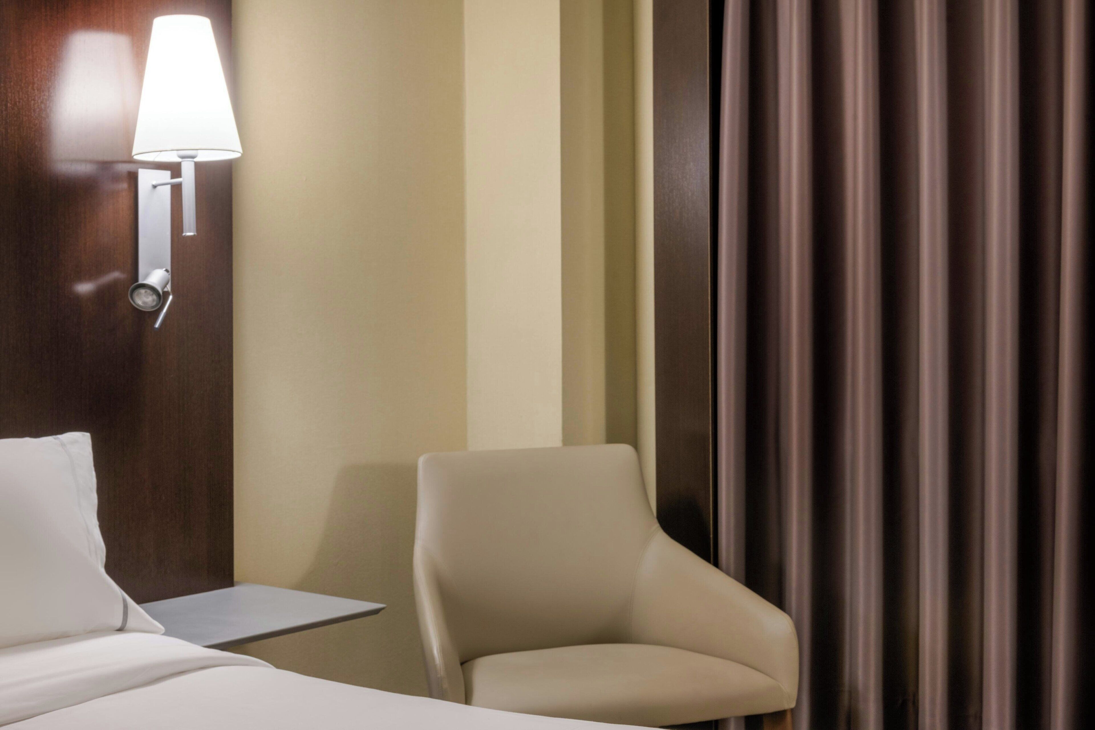 Una habitación de hotel con una cama, una silla, una mesita de noche, una lámpara y una cortina.