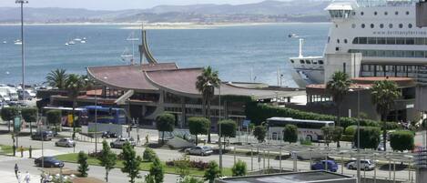 A coastal cityscape with a modern building featuring a glass facade, a bus stop, and a view of the sea.
