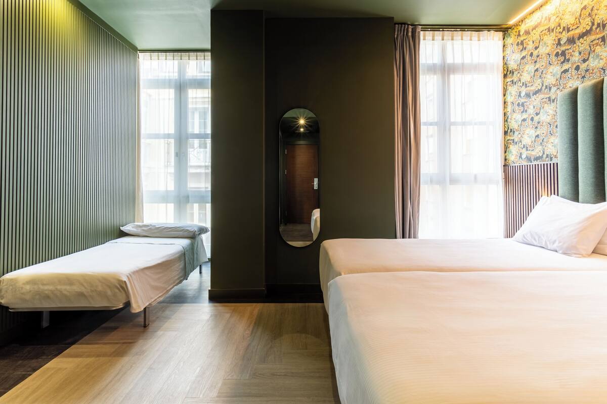 A hotel room with a large bed, a smaller bed, a mirror, and a window with curtains.