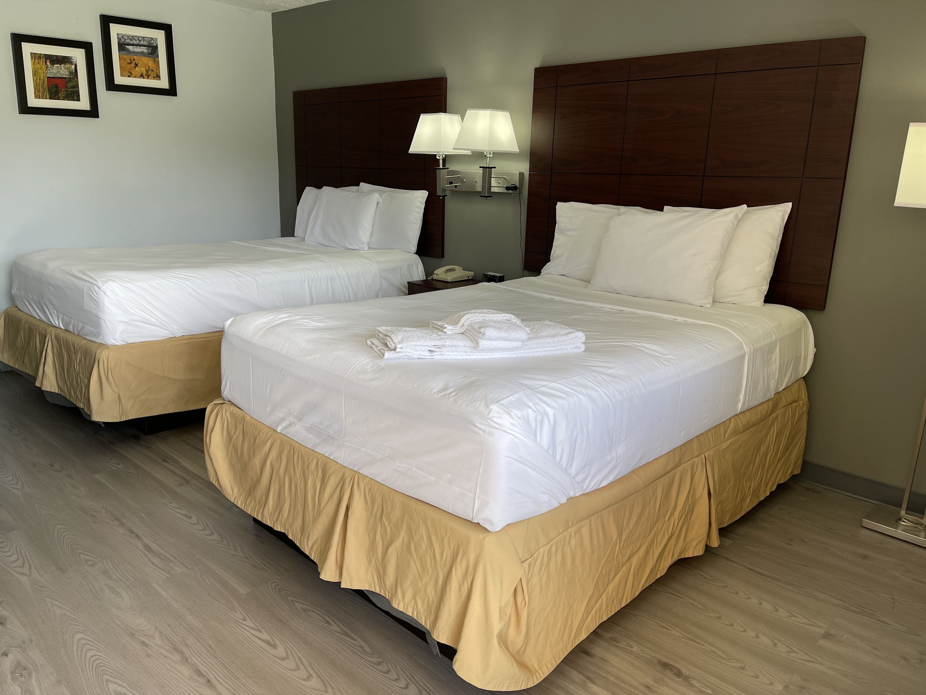 Une chambre d’hôtel avec deux lits, des couvre-lits beiges, une tête de lit en bois, une table de chevet avec une lampe et un tableau encadré au mur.
