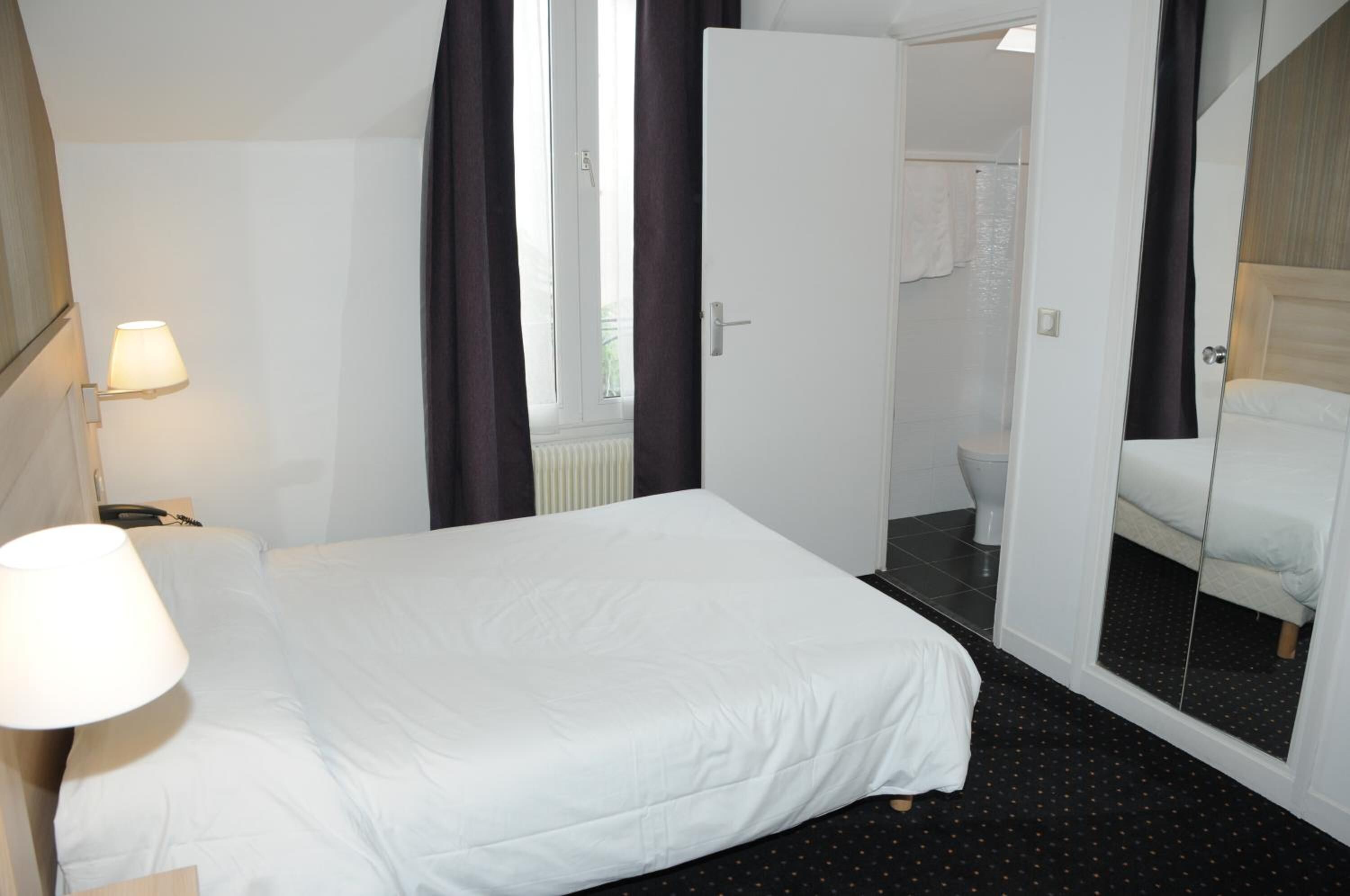 Habitación de hotel con cama, mesita de noche, lámpara, ventana con cortinas, puerta que da a un baño y un espejo que refleja la cama.
