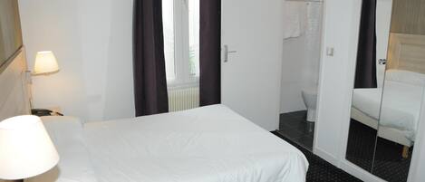 Habitación de hotel con cama, mesita de noche, lámpara, ventana con cortinas, puerta que da a un baño y un espejo que refleja la cama.