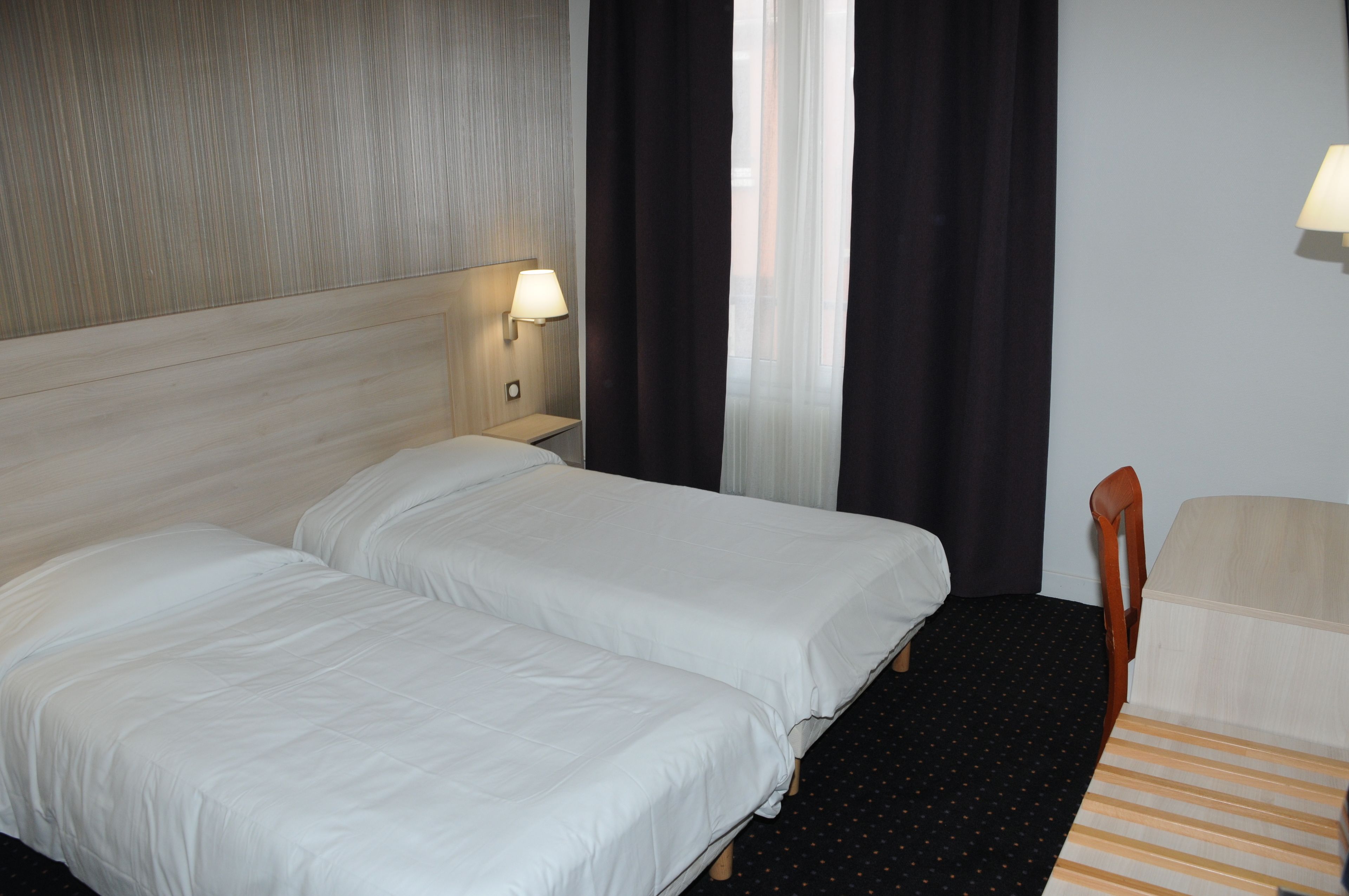 A hotel room with two single beds, a wooden headboard, a nightstand with a lamp, a chair, and a window with curtains.